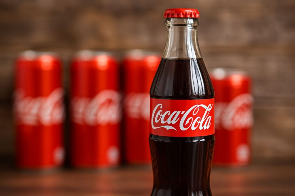 una bottiglia di Coca Cola con delle lattine di Coca Cola dietro