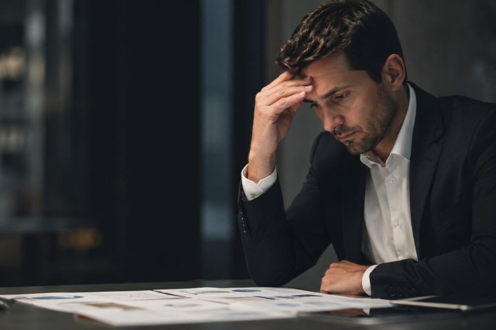 Uomo in ufficio, seduto al tavolo con documenti finanziari, mano sulla fronte, espressione preoccupata
