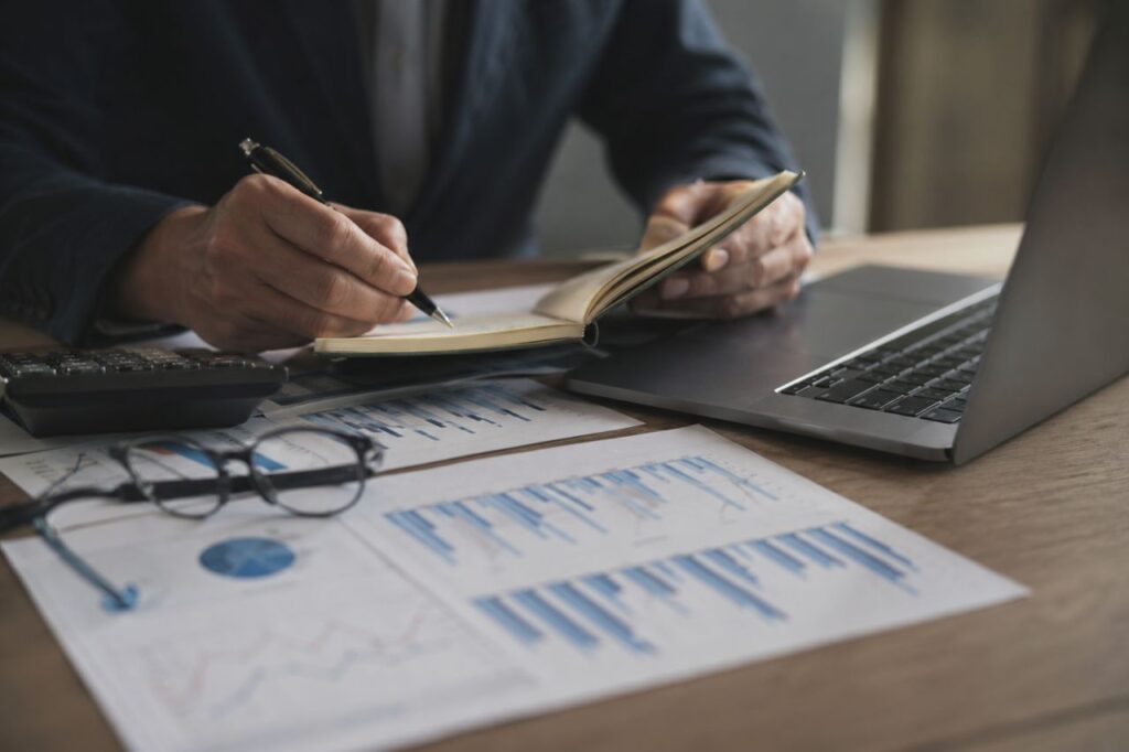 Mano di un professionista che scrive su un taccuino accanto a laptop, calcolatrice e grafici finanziari su scrivania.