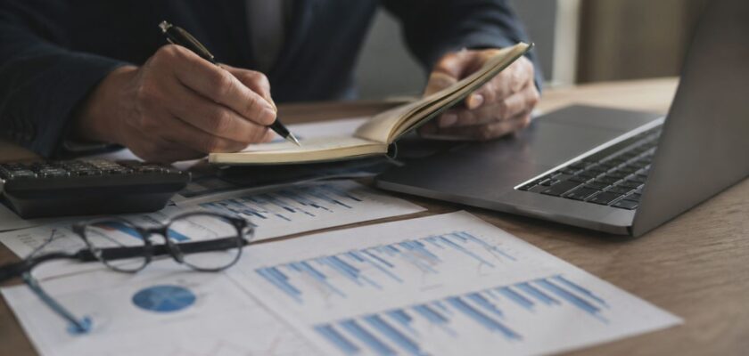 Mano di un professionista che scrive su un taccuino accanto a laptop, calcolatrice e grafici finanziari su scrivania.