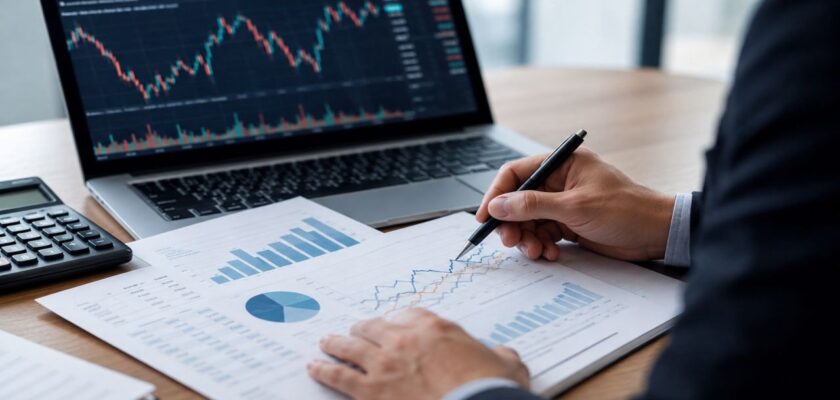 Scrivania da ufficio con laptop aperto su grafici finanziari, documenti analizzati a penna e calcolatrice accanto