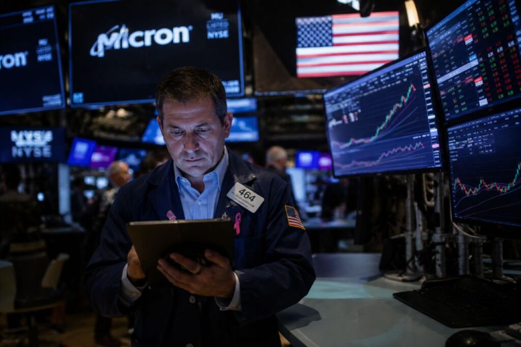Trader nella sala della Borsa di New York davanti a monitor finanziari con grafici azionari e logo Micron sullo sfondo