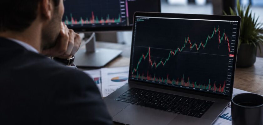 Trader seduto alla scrivania osserva grafici finanziari su laptop e monitor, in un ambiente professionale dedicato all'analisi dei mercati