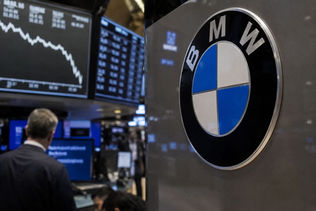 Logo BMW in primo piano davanti a monitor finanziari con grafici in calo, all'interno di una sala operativa di Borsa