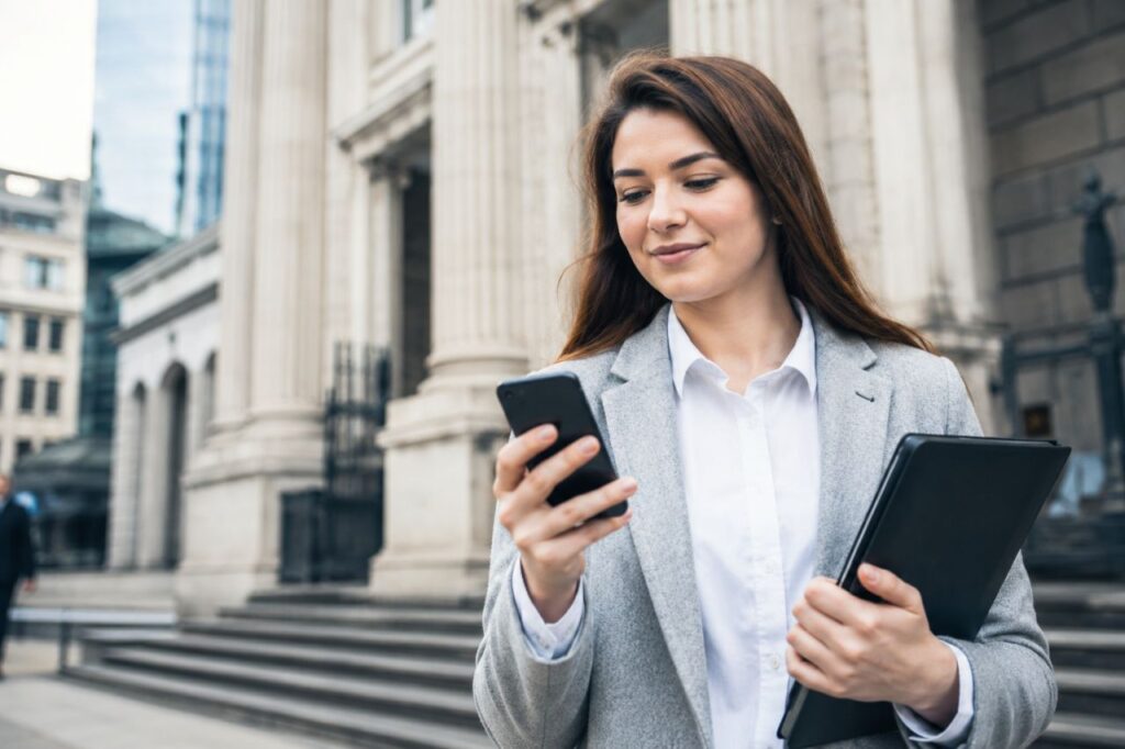 Giovane professionista consulta lo smartphone davanti a un edificio bancario, con tablet in mano e contesto urbano moderno