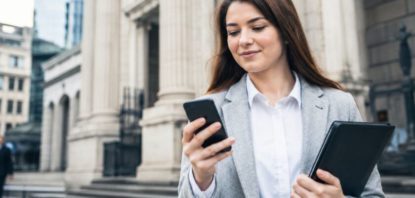 Giovane professionista consulta lo smartphone davanti a un edificio bancario, con tablet in mano e contesto urbano moderno