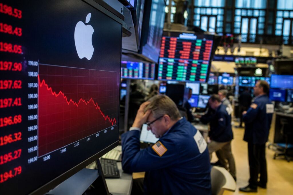 Operatore di Borsa davanti a monitor con logo Apple e grafico in calo, sullo sfondo una sala trading con schermi finanziari e altri trader al lavoro