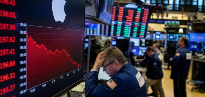 Operatore di Borsa davanti a monitor con logo Apple e grafico in calo, sullo sfondo una sala trading con schermi finanziari e altri trader al lavoro