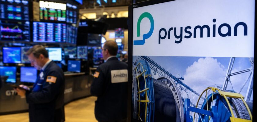 Schermo con il logo Prysmian e grandi bobine di cavi industriali in primo piano, sullo sfondo una sala operativa di borsa con trader e monitor finanziari