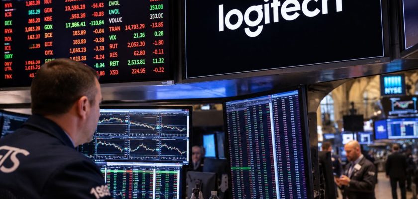 Trader davanti a monitor finanziari in una sala di Borsa, con display Logitech sullo sfondo e grafici di mercato