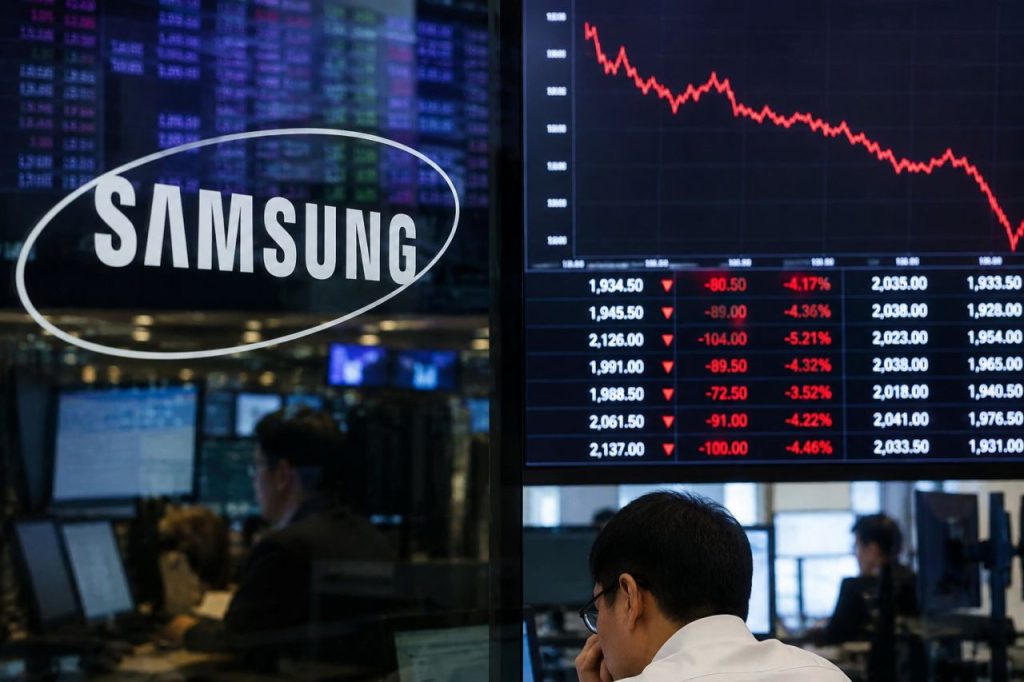 Logo Samsung accanto a monitor finanziari con grafico azionario in forte calo e operatori al lavoro
