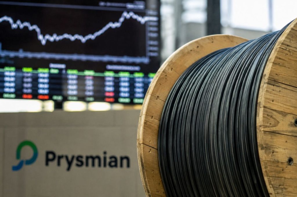 Bobina di cavi Prysmian in primo piano, con grafici e quotazioni di Borsa sullo sfondo