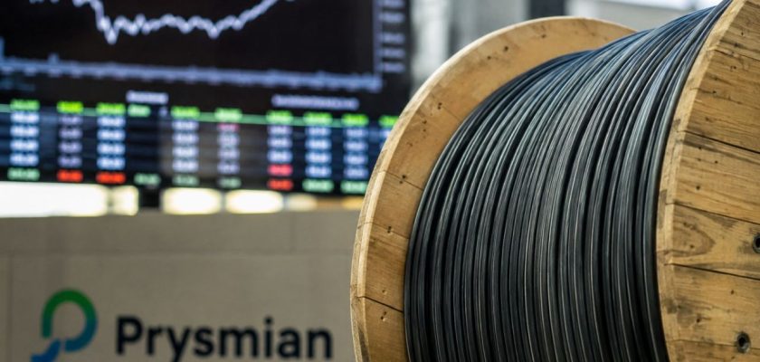 Bobina di cavi Prysmian in primo piano, con grafici e quotazioni di Borsa sullo sfondo