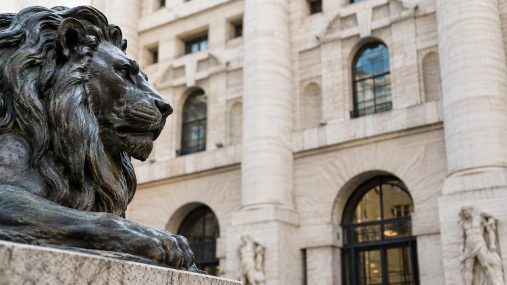 Statua di leone in bronzo in primo piano, davanti alla facciata monumentale di un edificio della Borsa con colonne e grandi archi